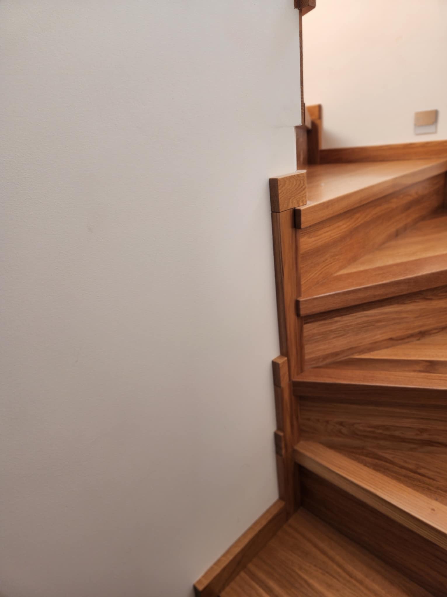 Drewniane schody spiralne, widok z dołu, z bliska, z jasną ścianą w tle. Widoczne detale wykończenia, naturalny odcień drewna, elegancki minimalizm.