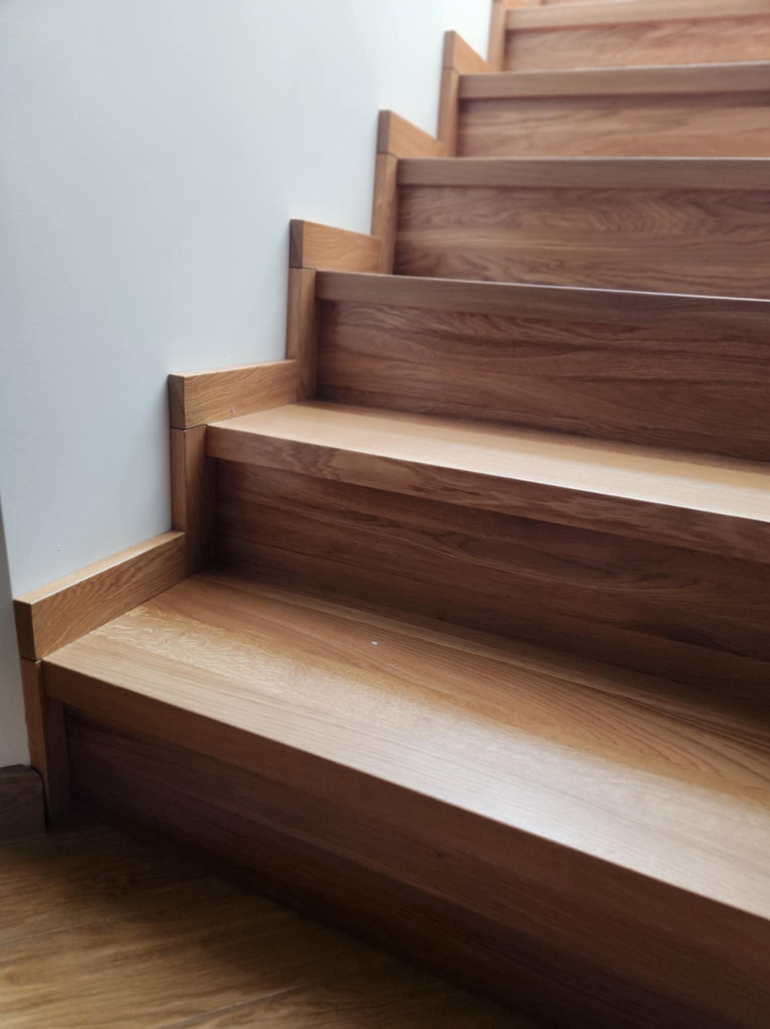 Detal drewnianych schodów dębowych z bliska, widoczne usłojenie i subtelne oświetlenie. Minimalistyczny design, jasna ściana w tle.