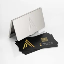 Stylowe czarne karty z błyszczącym złotym logo i kodem QR, ułożone obok otwartego metalowego wizytownika z wytłoczonym logo na białym tle.