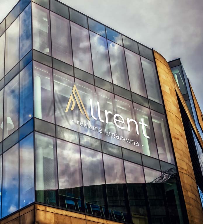 Zewnętrzna reklama firmy 'Allurent - reklama kreatywna' umieszczona na szklanej fasadzie nowoczesnego budynku biurowego, z odbiciem chmur i nieba w szybach.