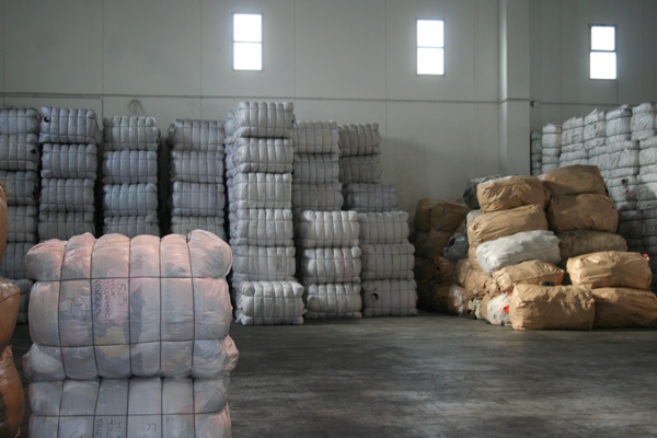 Magazyn z paletami sprasowanej odzieży używanej, część w folii, część w papierowych workach, widoczne napisy na folii i workach.