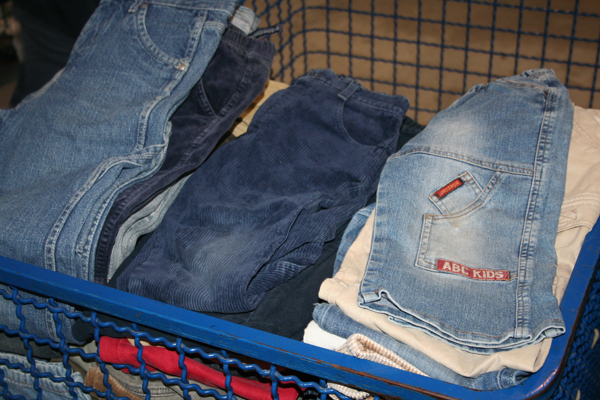 Kosz z niebieskiej metalowej siatki wypełniony stertą używanej odzieży, głównie spodni jeansowych, sztruksowych i beżowych, z widocznym czerwonym elementem na dnie kosza.