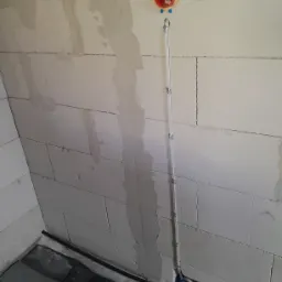 Instalacja elektryczna w trakcie montażu w surowym wnętrzu: pomarańczowa puszka z przewodami na ścianie z betonu komórkowego, pionowy peszel przymocowany metalowymi uchwytami, czarne kable...