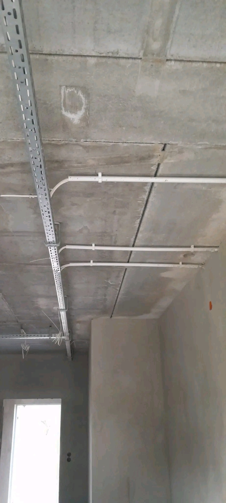 Szara, surowa przestrzeń z widocznymi, zamontowanymi kanałami i rurami instalacji pod sufitem. Widoczne niedoskonałości betonu i surowy stan wykończenia wnętrza.