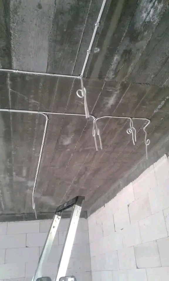 Szare, surowe wnętrze z widoczną instalacją elektryczną na suficie i drabiną opartą o ścianę z białych bloczków. Kable zwisają luźno, czekając na podłączenie.