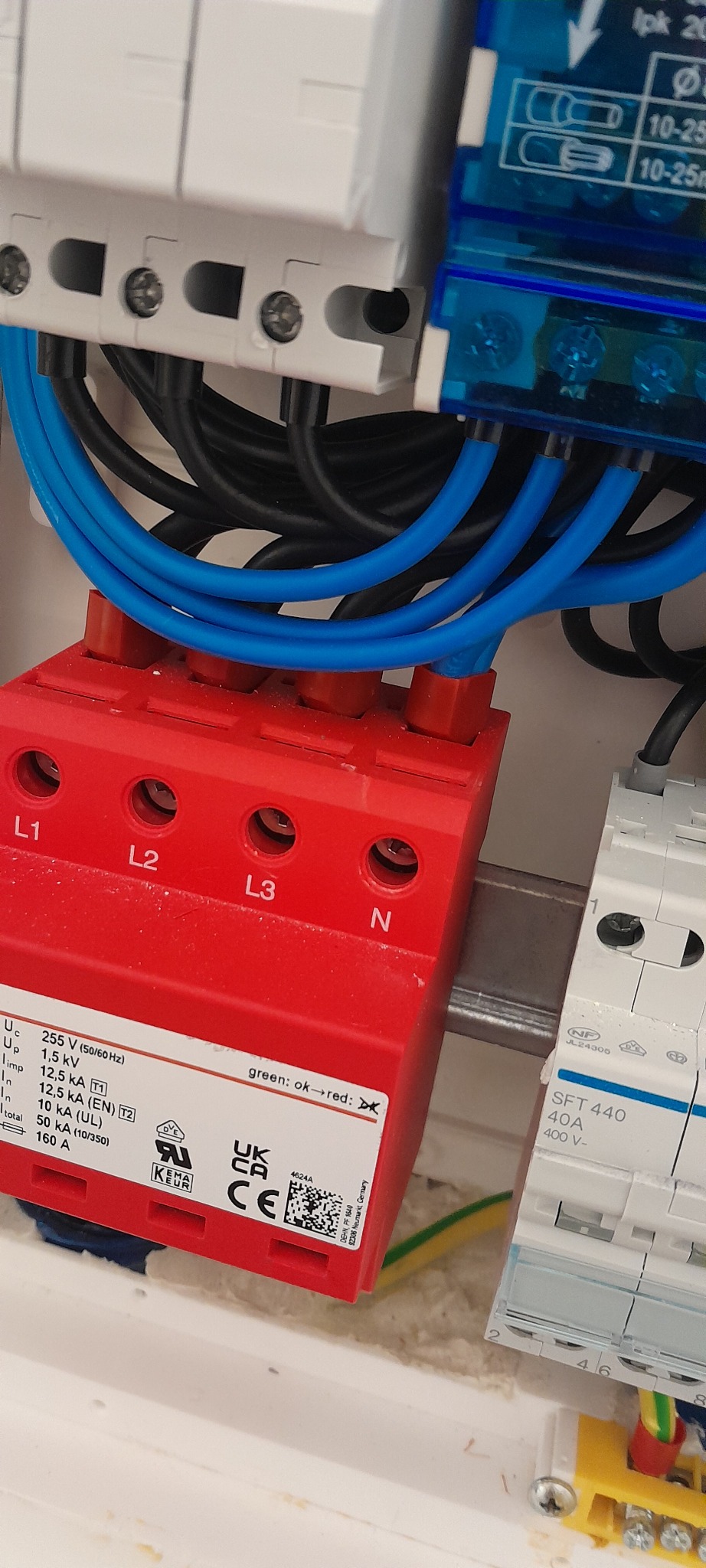 Zabezpieczenie przeciwprzepięciowe w rozdzielnicy, z podłączonymi przewodami w kolorach niebieskim i czarnym. Widoczne oznaczenia L1, L2, L3, N na czerwonym module.