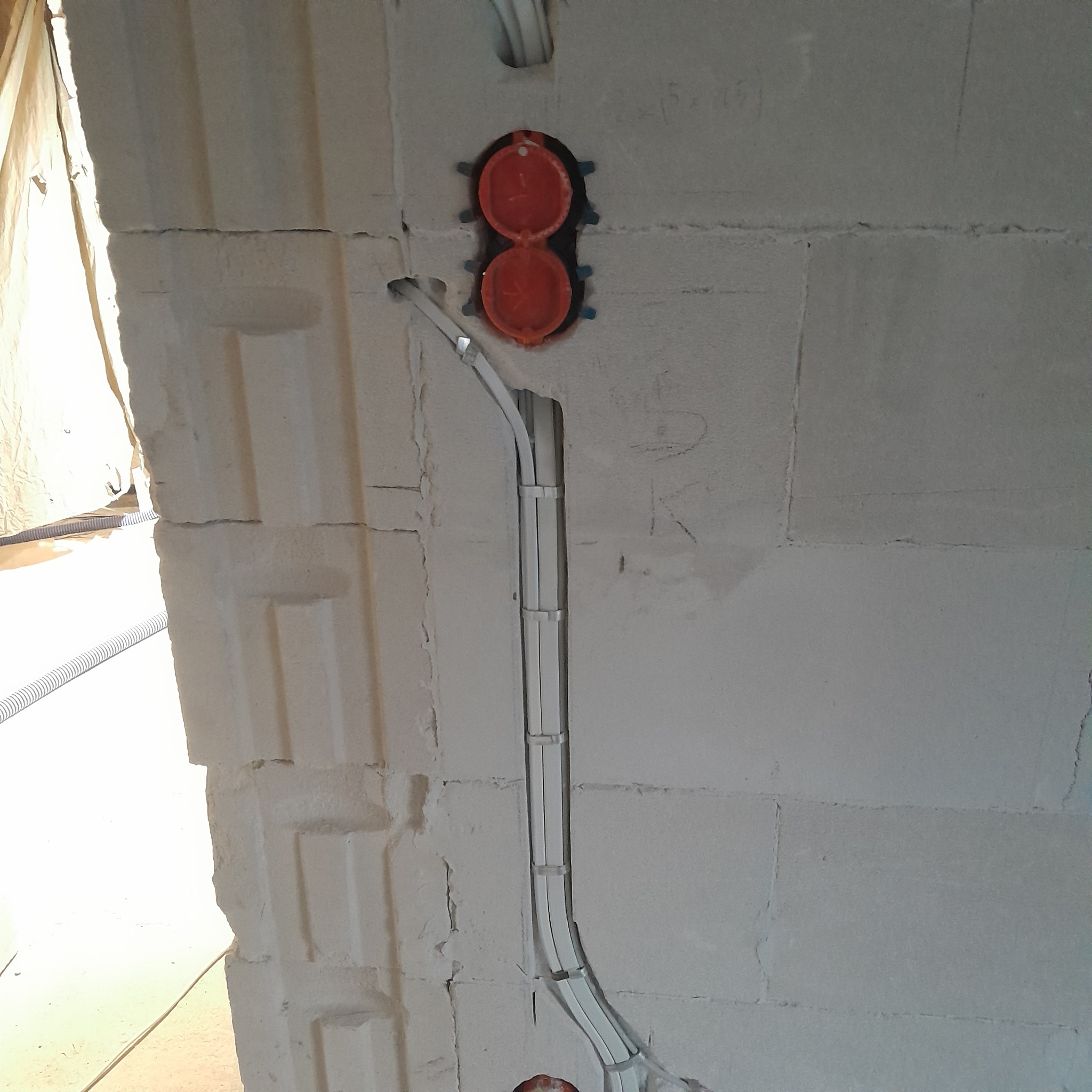 Białe kable w peszlu przymocowane do ściany z betonu komórkowego, widoczne pomarańczowe puszki podtynkowe. Etap prac w nowym budownictwie.