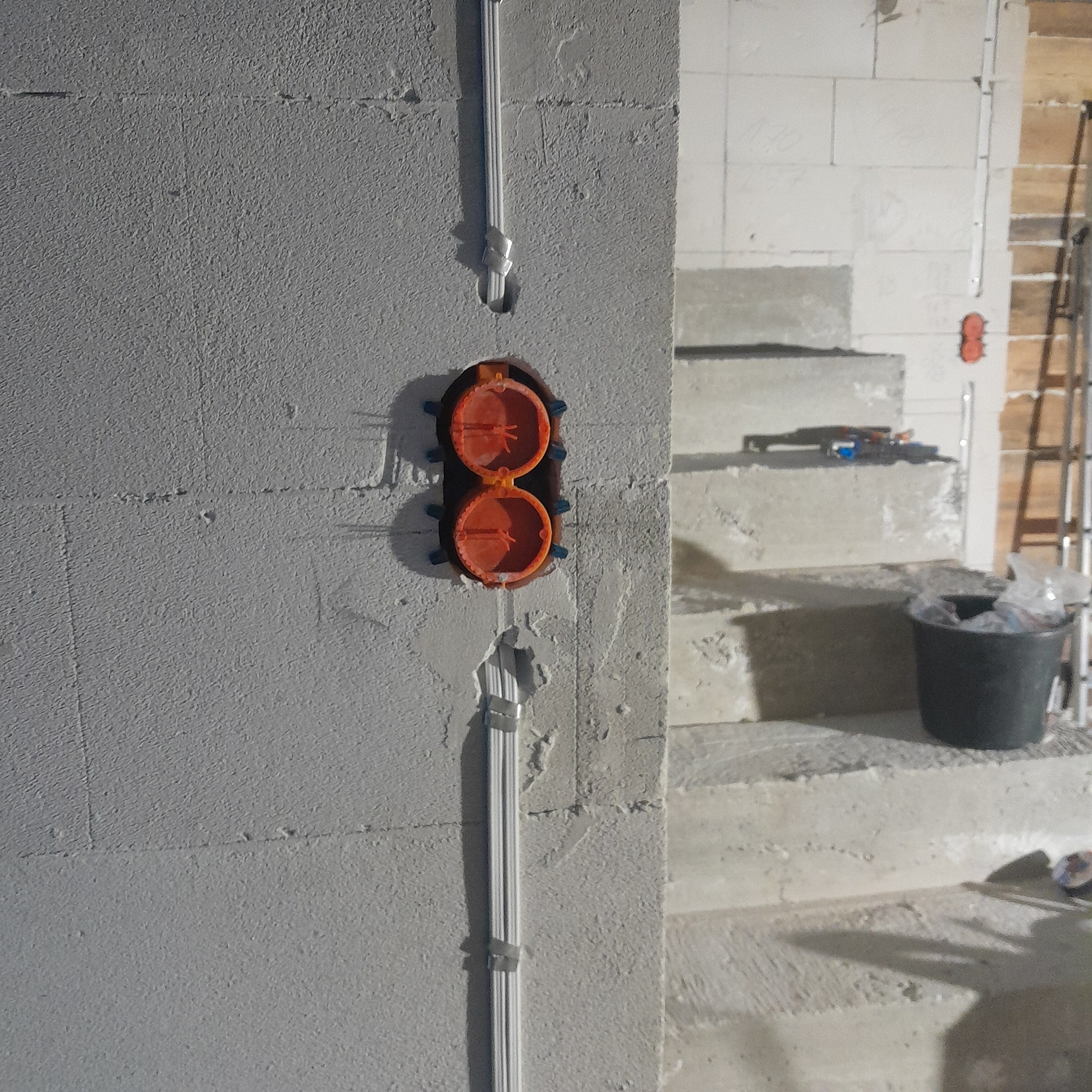 Montaż podwójnej, pomarańczowej puszki elektrycznej w szarej, surowej ścianie z białymi przewodami. Widoczne schody i drabina w tle. Etap prac instalacyjnych.