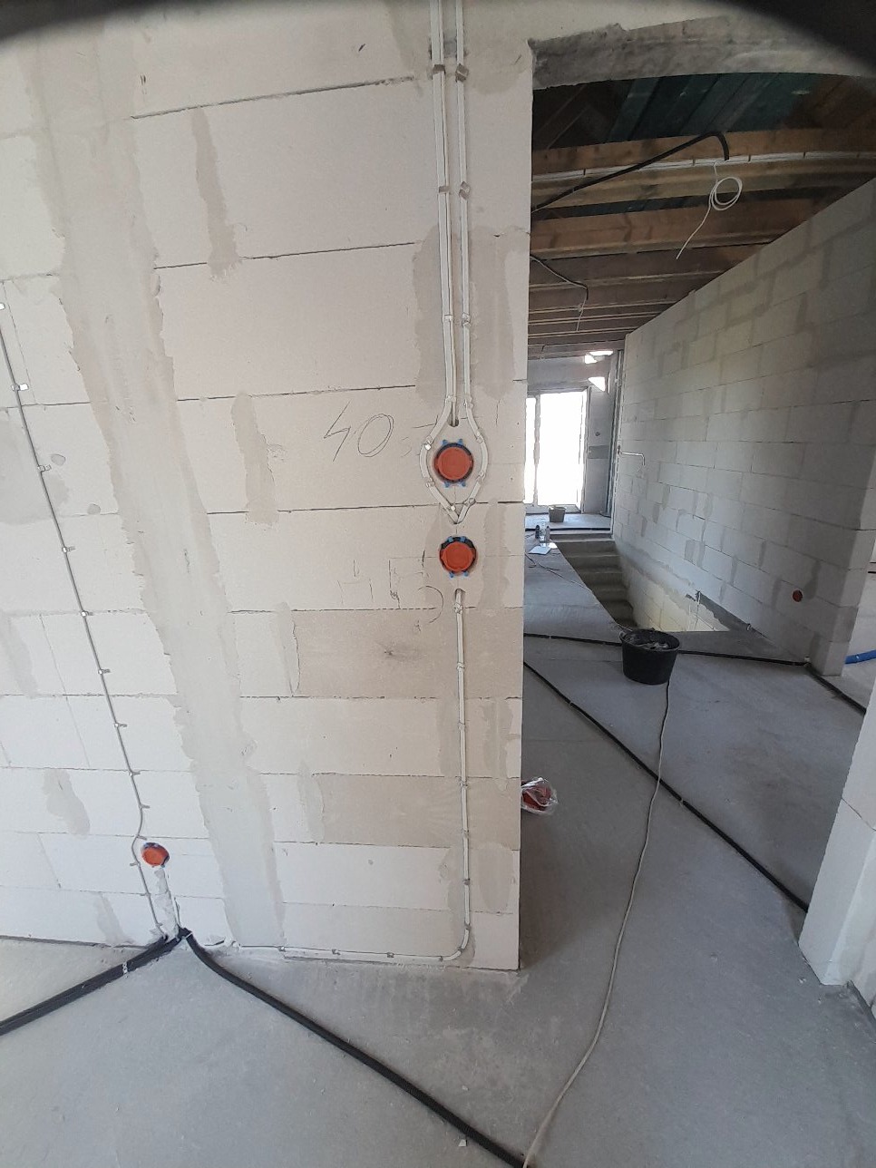 Białe ściany z instalacją przewodów i puszek elektrycznych w surowym wnętrzu budynku w trakcie budowy. Widoczne kable na podłodze i elementy konstrukcji stropu.