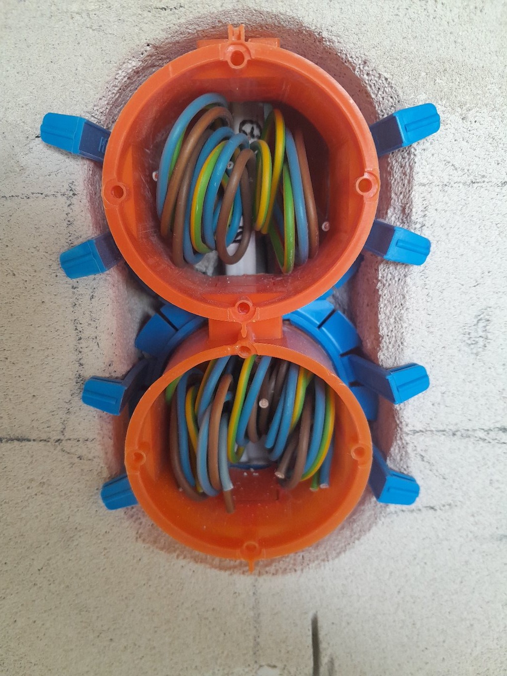 Dwa pomarańczowe gniazdka elektryczne z niebieskimi mocowaniami, wypełnione kolorowymi przewodami, osadzone w surowej ścianie. Widok z bliska.