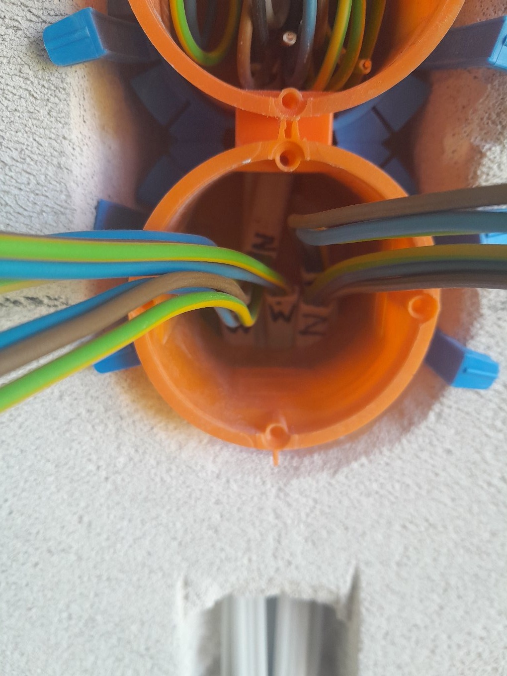 Pomarańczowa puszka instalacyjna z niebieskimi elementami mocującymi, z widocznymi przewodami wewnątrz, osadzona w białej ścianie. Ujęcie z bliska, z góry.