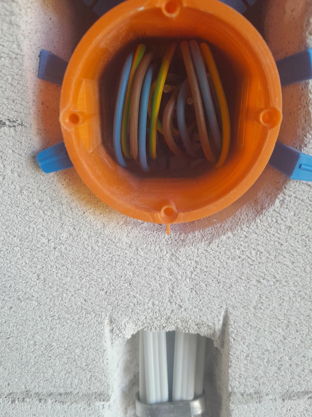 Pomarańczowa puszka instalacyjna z kolorowymi przewodami w szarej ścianie. Widoczne białe rurki instalacyjne w dolnej części kadru.