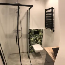 Nowoczesna łazienka z kabiną prysznicową typu walk-in z czarnymi ramami, wiszącą toaletą na tle ściany z motywem tropikalnych liści oraz czarnym grzejnikiem drabinkowym.