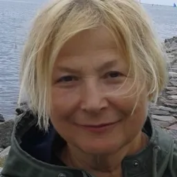 Portret uśmiechniętej kobiety z blond włosami w zielonej kurtce na tle morza z żaglówkami.