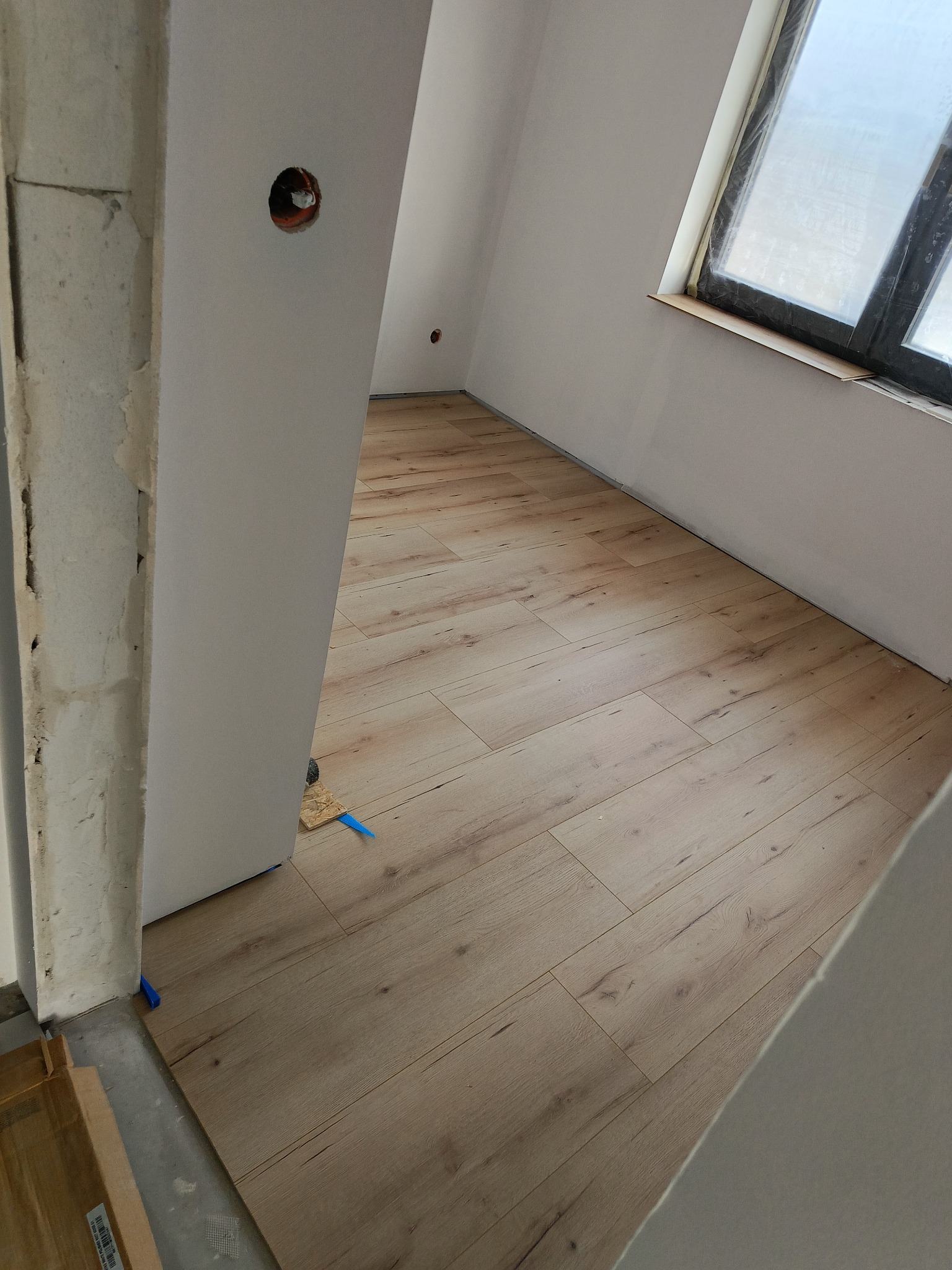 Jasne panele podłogowe ułożone w pokoju na poddaszu, widoczne fragmenty ściany z oknem dachowym i surowego otworu drzwiowego. Kliny dystansowe przy ścianie.