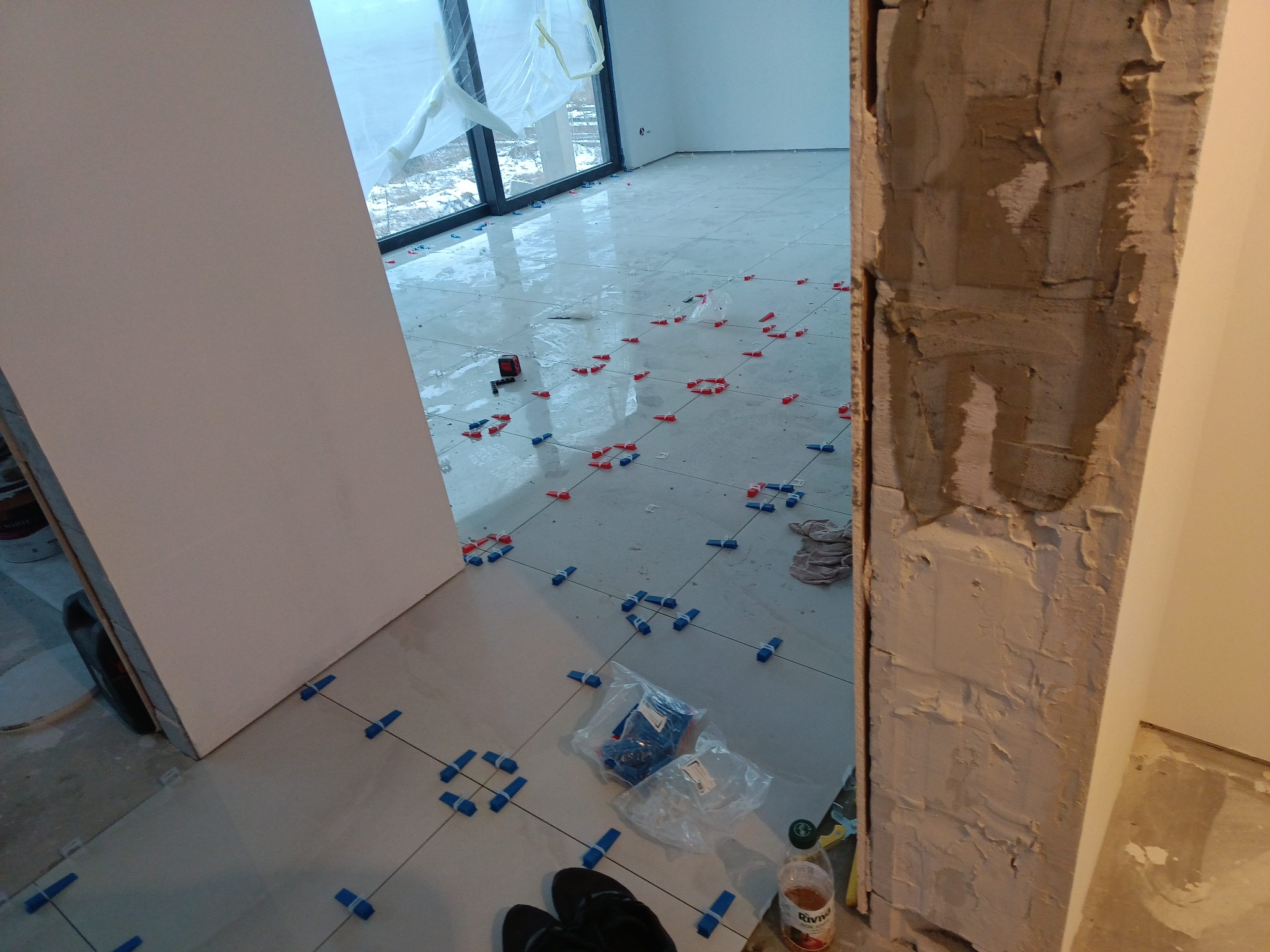 Układanie jasnych płytek podłogowych z użyciem klipsów poziomujących. Widoczne narzędzia i materiały budowlane. Duże okno w tle. Prace wykończeniowe w trakcie.