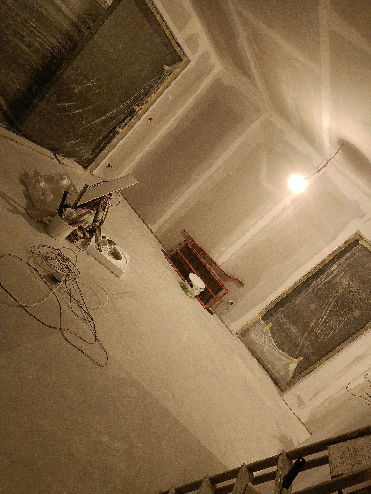 Skośny kadr wnętrza w trakcie prac wykończeniowych: ściany i sufit pokryte gładzią, zabezpieczone okna, narzędzia pracy, drabina. Widoczne kable i lampa oświetlająca.