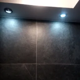 Ściana wyłożona ciemnymi, matowymi płytkami w łazience, oświetlona trzema punktowymi lampami sufitowymi, z których jedna jest reflektorem.