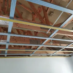 Konstrukcja sufitu podwieszanego z profili metalowych, widoczne drewniane elementy więźby dachowej i instalacja elektryczna, przygotowanie do montażu płyt gipsowo-kartonowych.