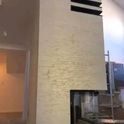 Kominek obudowany pionową ścianą z efektem dekoracyjnym w odcieniach beżu i szarości, z czarnymi, poziomymi wnękami w górnej części i czarnym wkładem kominkowym na dole, widoczna drabina w tle.