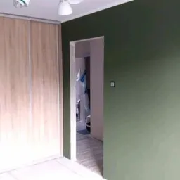 Świeżo pomalowane na oliwkowy kolor ściany w pokoju z przesuwną szafą w jasnym odcieniu drewna, widok na sąsiednie pomieszczenie z widocznymi elementami wyposażenia.