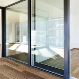 Nowoczesne przesuwne okna balkonowe z czarnymi ramami, widok z drewnianego tarasu na jasne wnętrze z kuchnią i parkietem.