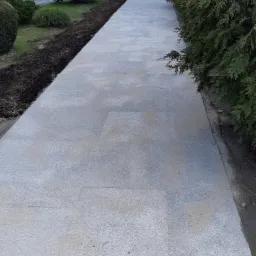 Świeżo ułożony chodnik z jasnoszarej kostki granitowej, obrzeża z ciemniejszej kostki, widoczne przygotowane podłoże pod trawnik i rząd tuj, pomarańczowy pachołek na pierwszym planie.