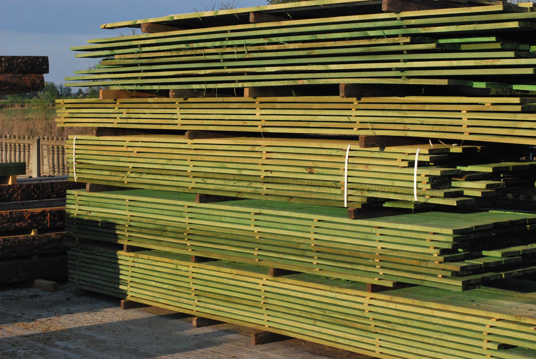 Stosy impregnowanej, zielonej tarcicy budowlanej ułożone na przekładkach, z widocznym fragmentem ogrodzenia w tle.