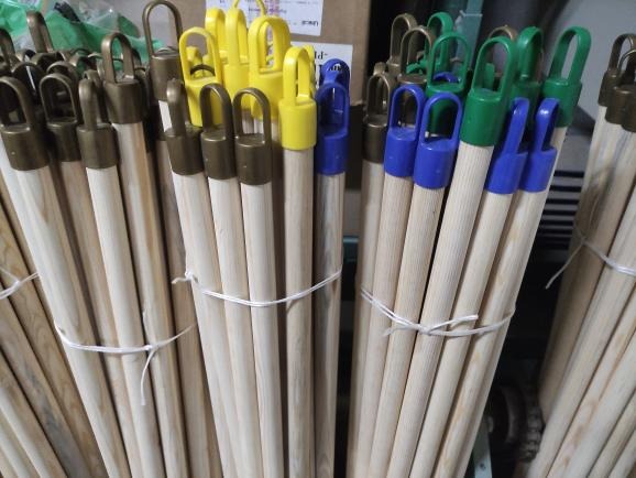 Pęczki drewnianych trzonków do narzędzi z kolorowymi plastikowymi uchwytami (brązowe, żółte, niebieskie, zielone) powiązane białym sznurkiem, widoczne w magazynie.