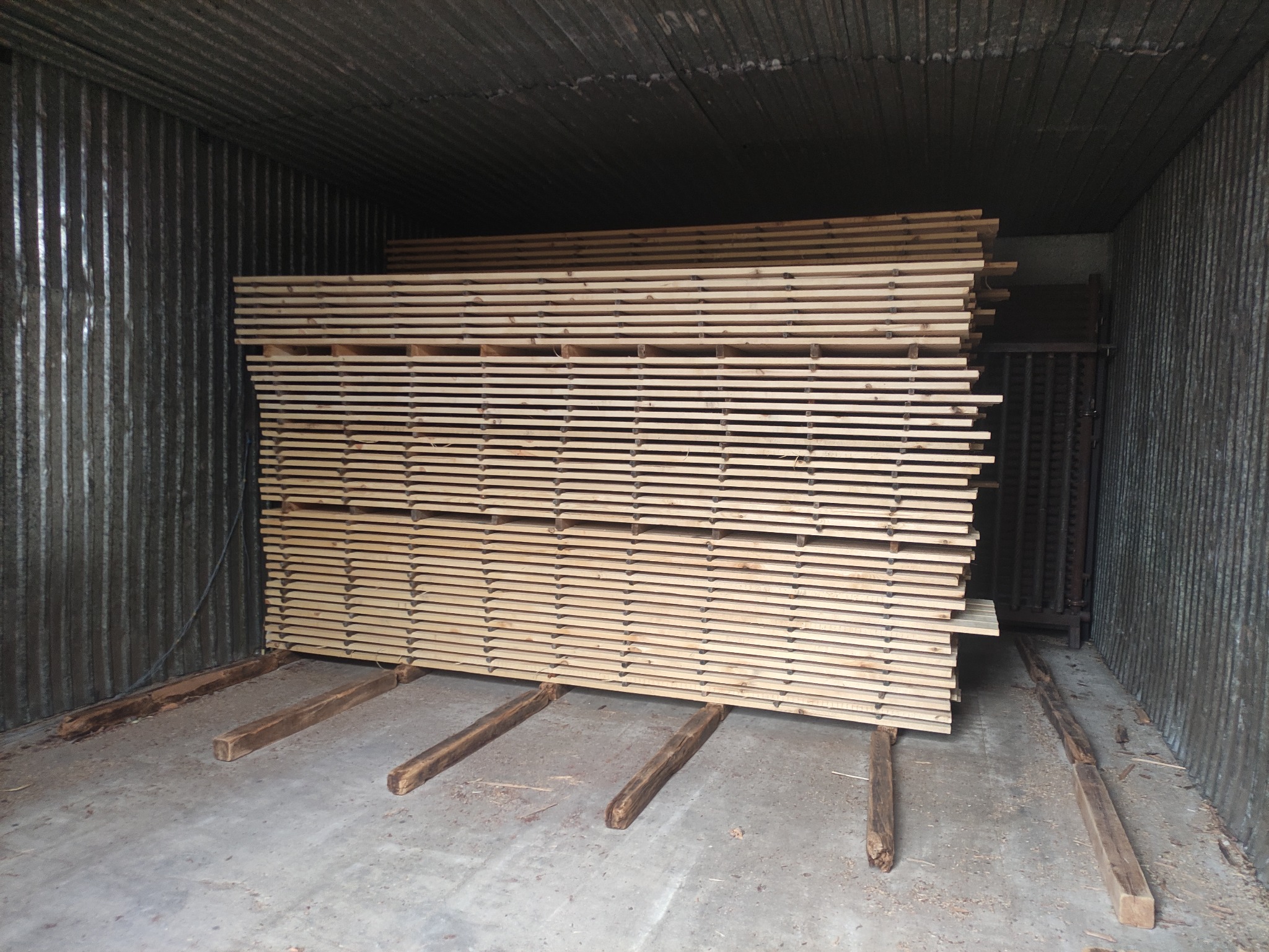 Stos tarcicy ułożony na drewnianych legarach w metalowym magazynie o ścianach z blachy falistej.