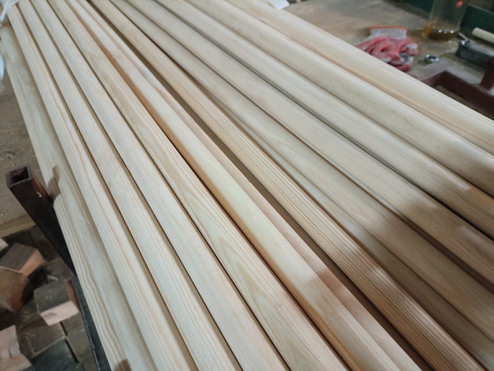 Stos równo ułożonych, jasnych, drewnianych listew o okrągłym przekroju, widoczne słoje drewna, w tle fragment warsztatu z rękawicami roboczymi.
