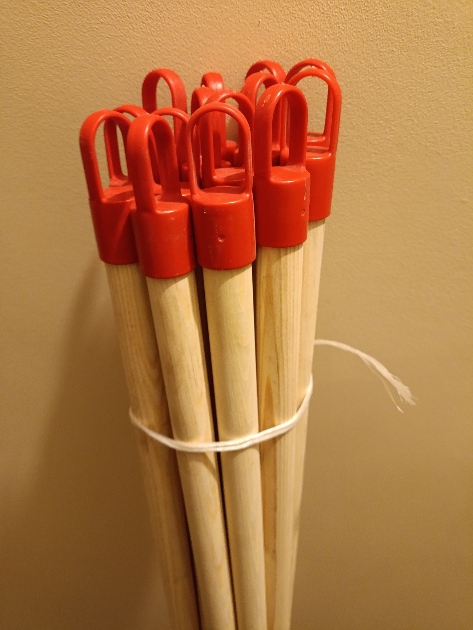 Wiązka drewnianych trzonków do narzędzi z czerwonymi plastikowymi uchwytami, związana sznurkiem na tle beżowej ściany.