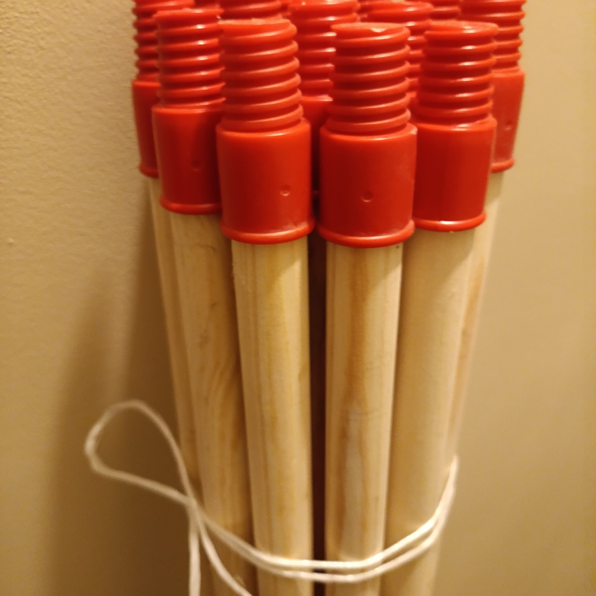 Wiązka jasnych, drewnianych trzonków do narzędzi z czerwonymi, plastikowymi nakrętkami na końcach, związana białym sznurkiem, na tle beżowej ściany.