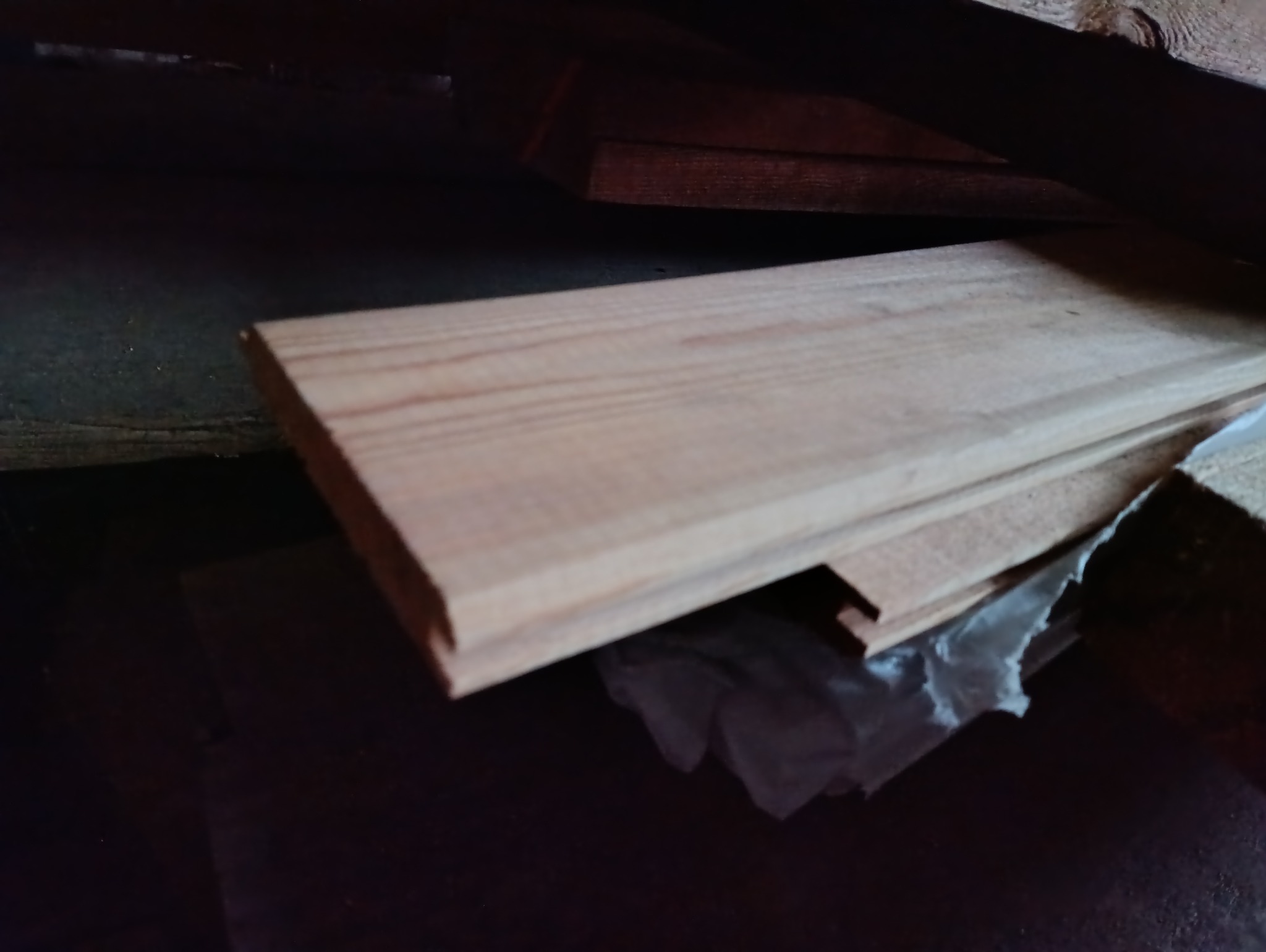 Stos jasnych desek z widocznym usłojeniem, częściowo przykrytych folią, w słabo oświetlonym pomieszczeniu o drewnianej konstrukcji.