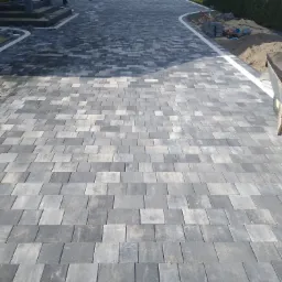 Ułożona szara kostka na podjeździe, z widocznym fragmentem domu w tle i osobą przechodzącą obok.