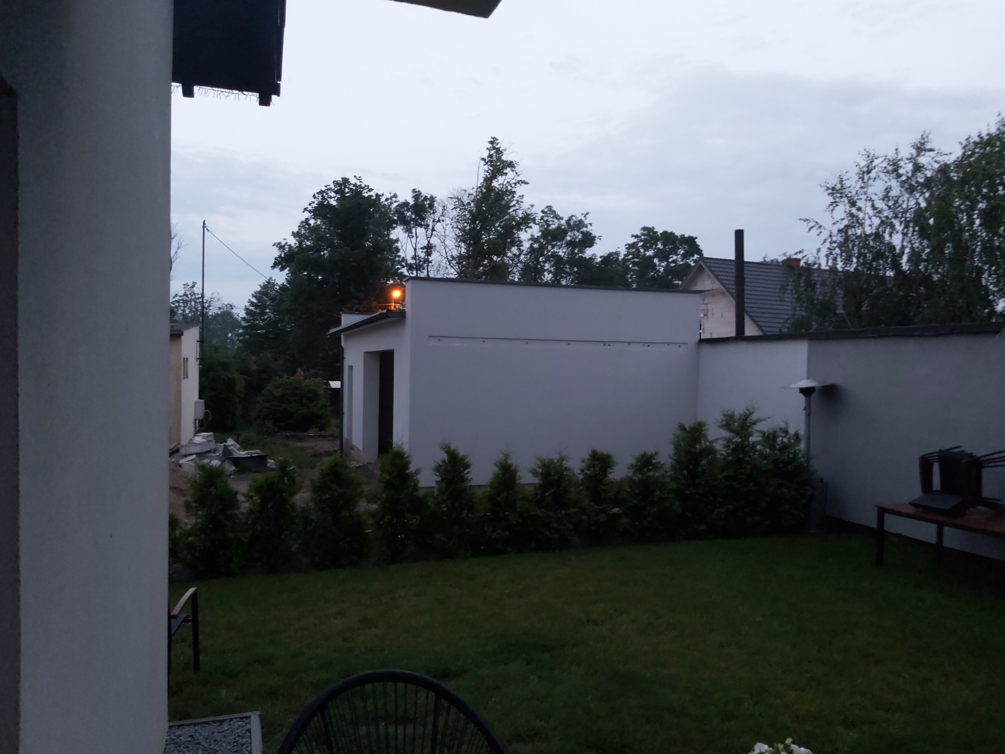 Biały budynek gospodarczy z oświetleniem zewnętrznym i rzędem małych drzew iglastych na tle trawnika i zachmurzonego nieba o zmierzchu.