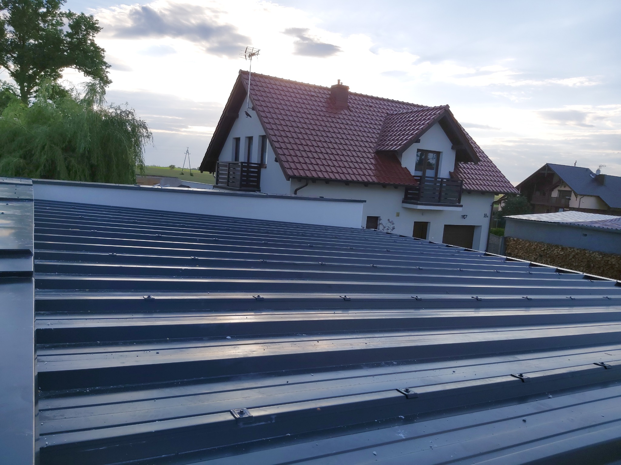 Nowoczesne pokrycie dachu z blachy trapezowej w kolorze grafitowym, widok z góry, w tle dom jednorodzinny z czerwoną dachówką i białymi ścianami w słoneczny dzień.