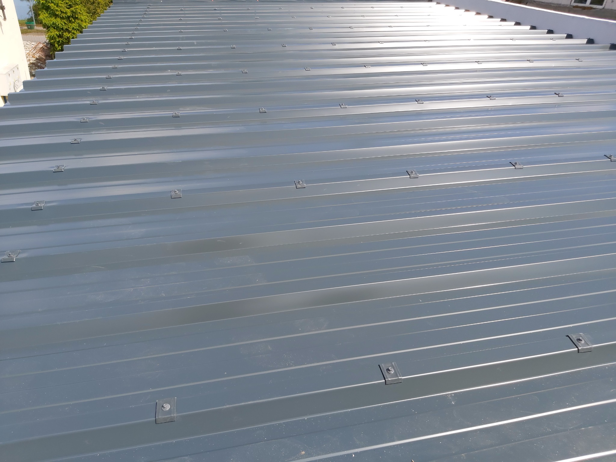 Nowe, szare blaszane pokrycie dachowe z widocznymi mocowaniami, ułożone równolegle, z fragmentem zieleni i białej ściany w tle.