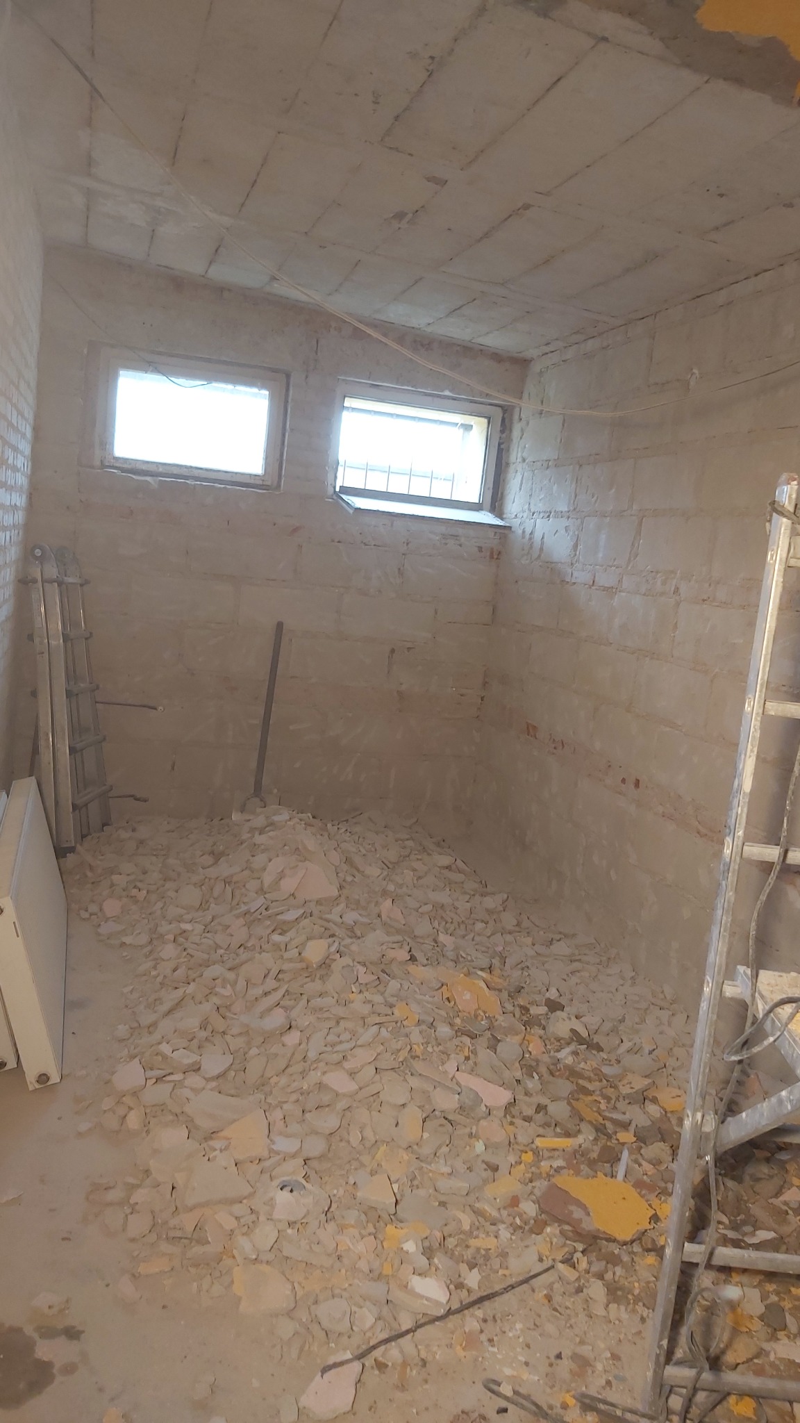 Wnętrze pomieszczenia w trakcie prac remontowych, widoczne fragmenty gruzu, odkryta cegła na ścianach, drabina oparta o ścianę, dwa małe okna.