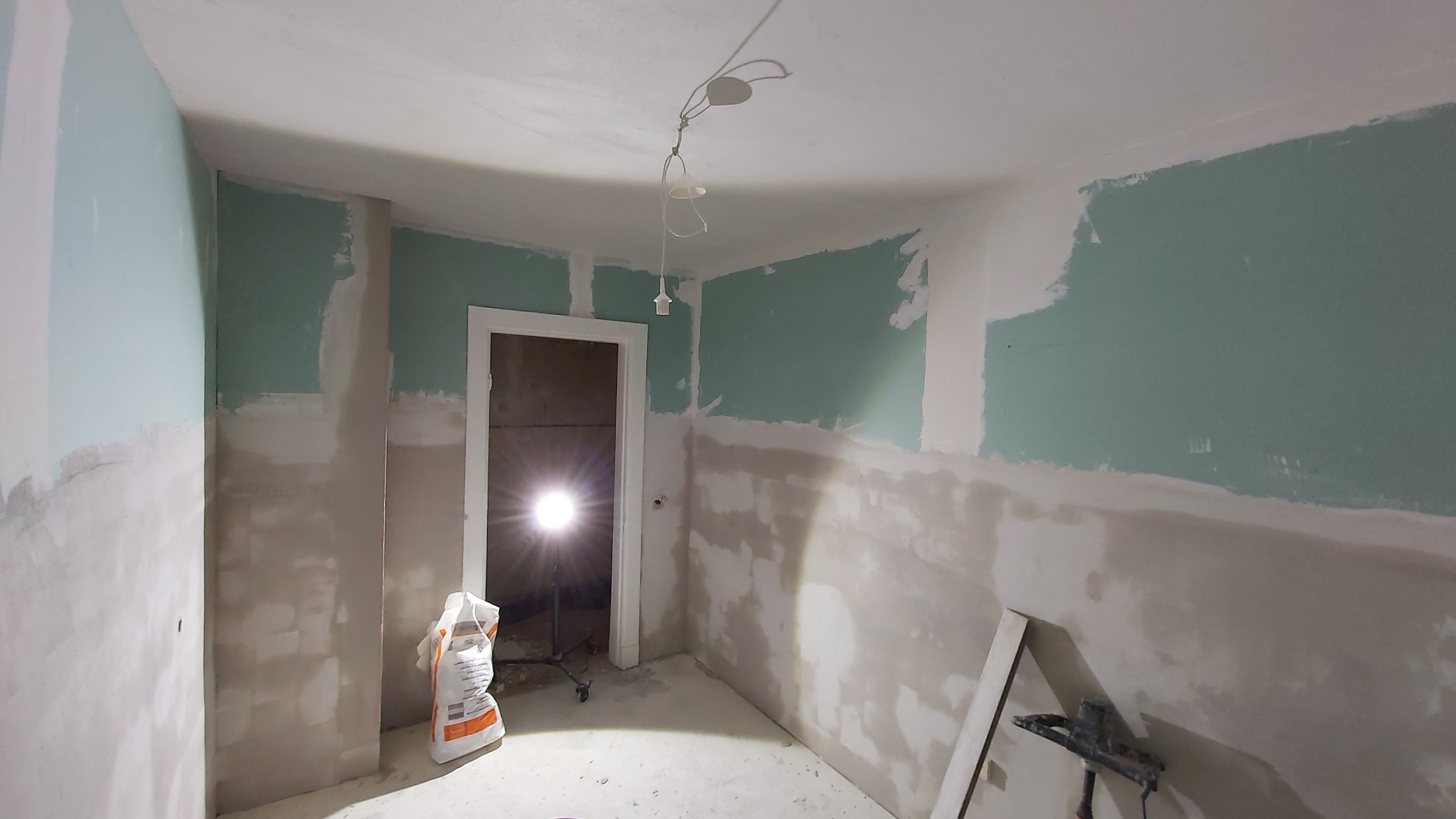 Wnętrze w Nakle nad Notecią w trakcie prac wykończeniowych: ściany częściowo pokryte szarą gładzią, górna część ścian pomalowana na zielono, widoczny worek z zaprawą i lampa halogenowa.