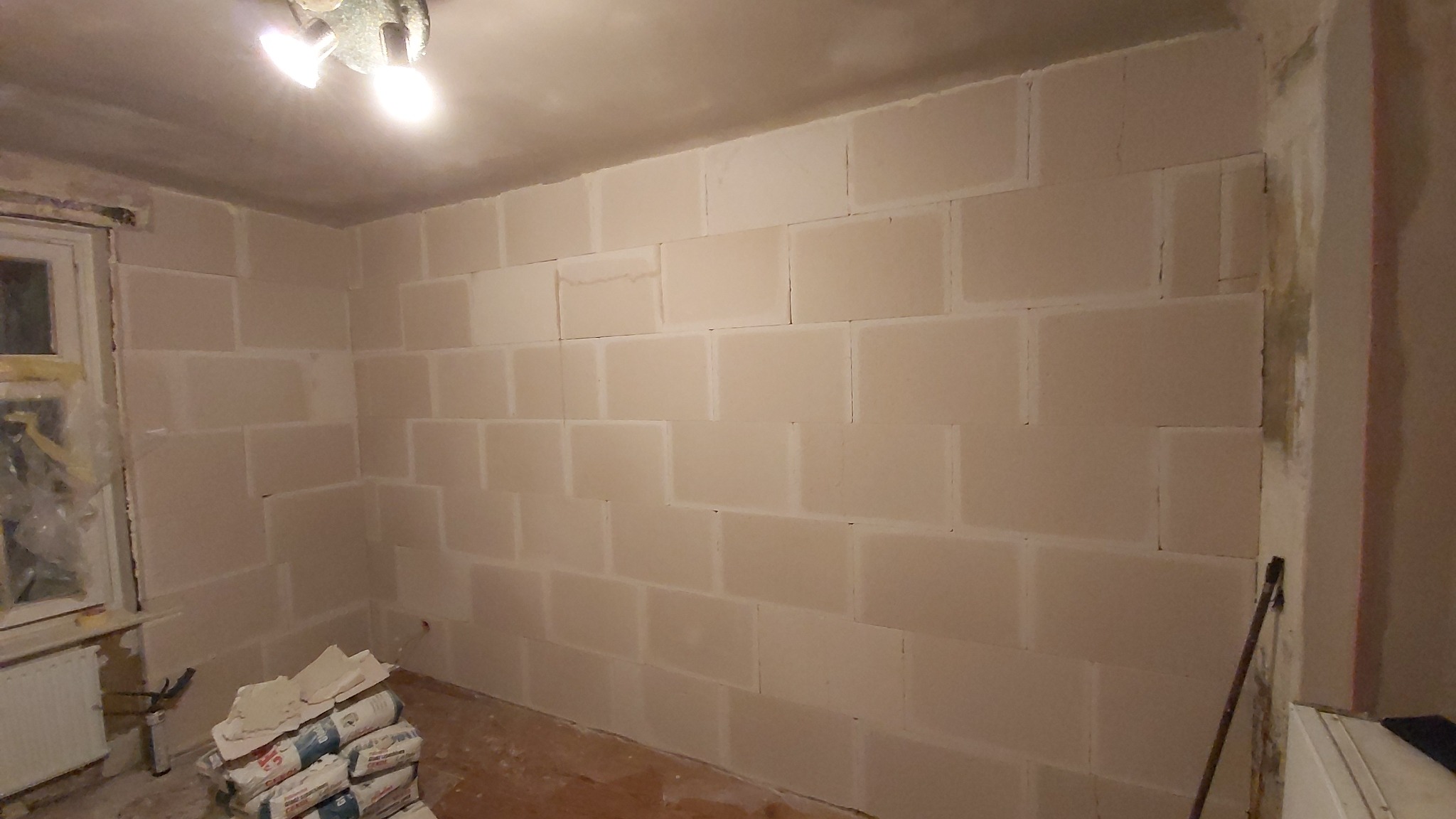 Wnętrze pokoju w trakcie remontu, ściany pokryte wstępnie gładzią gipsową układaną w kwadratowe segmenty, worki z gładzią przy ścianie, widoczne niedoskonałości wykończenia i oświetlenie sufitowe.