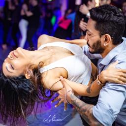 Anna Mazur Pracownia Wizerunku - Para w dynamicznej pozie tanecznej na parkiecie, kobieta odchylona do tyłu, mężczyzna w okularach ją podtrzymuje, w tle rozmazani tancerze i kolorowe światła.