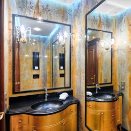 Anna Mazur Pracownia Wizerunku - Luksusowa łazienka z marmurowymi ścianami, złotą szafką pod umywalką z czarnym blatem, ozdobnym lustrem i kryształowym kinkietem.