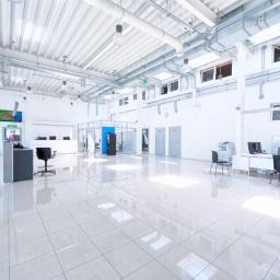 Anna Mazur Pracownia Wizerunku - Jasne, przestronne biuro z błyszczącą podłogą, widoczne stanowiska pracy, system wentylacji pod sufitem i roślina doniczkowa w oknie.