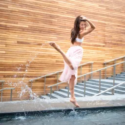 Kobieta w białym topie i różowej spódnicy tańczy na tle drewnianej ściany, unosząc nogę i rozpryskując wodę z fontanny.