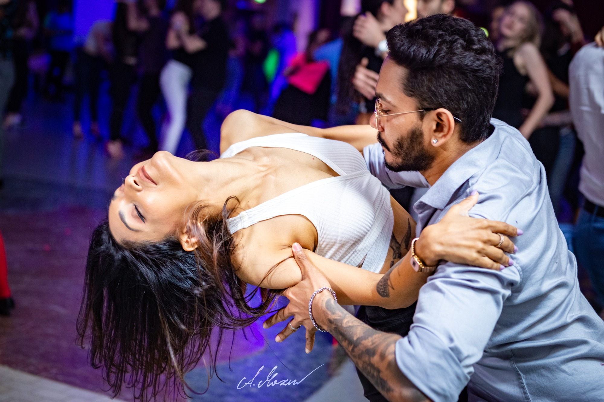 Para w dynamicznej pozie tanecznej na parkiecie, kobieta odchylona do tyłu, mężczyzna w okularach ją podtrzymuje, w tle rozmazani tancerze i kolorowe światła.