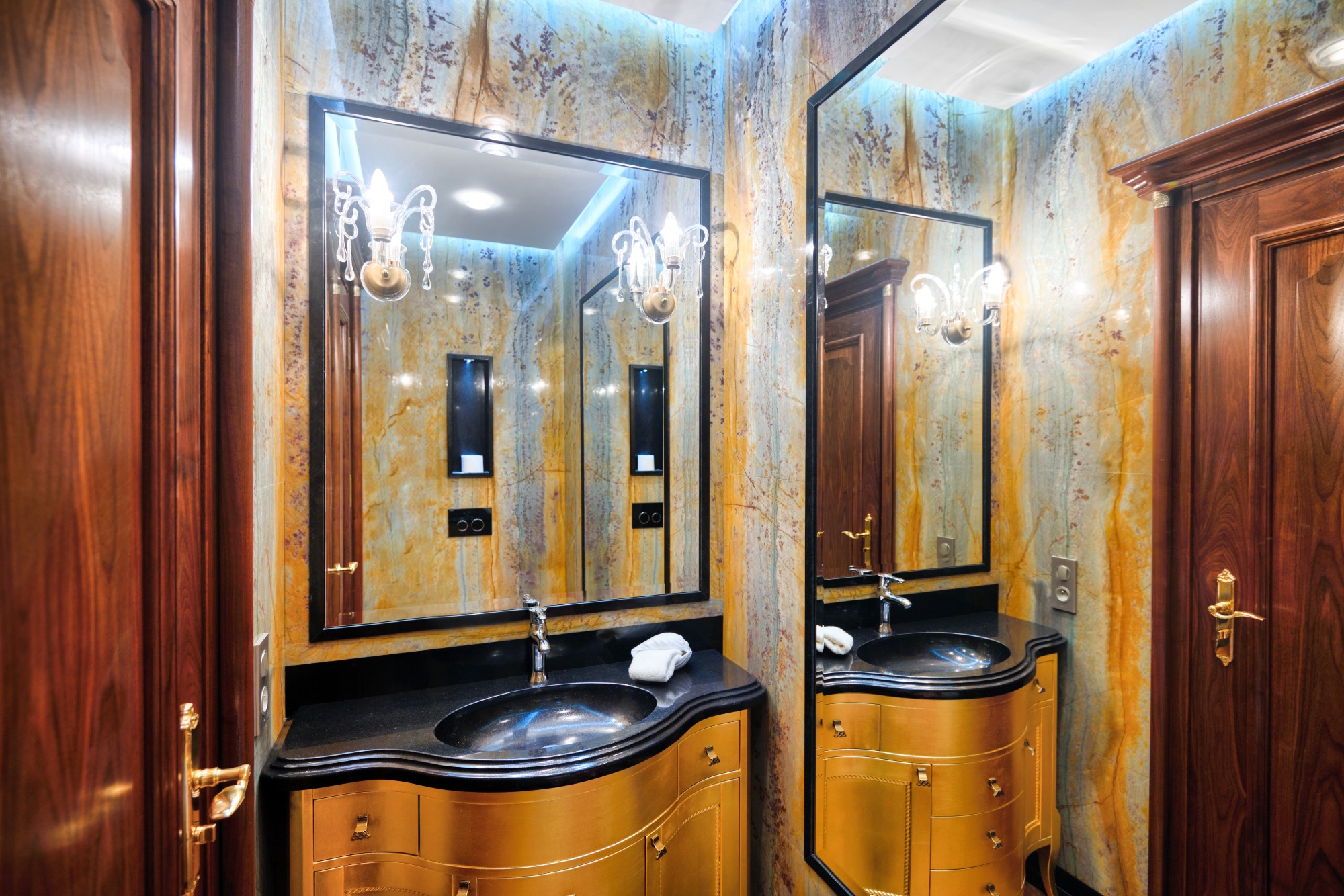 Luksusowa łazienka z marmurowymi ścianami, złotą szafką pod umywalką z czarnym blatem, ozdobnym lustrem i kryształowym kinkietem.