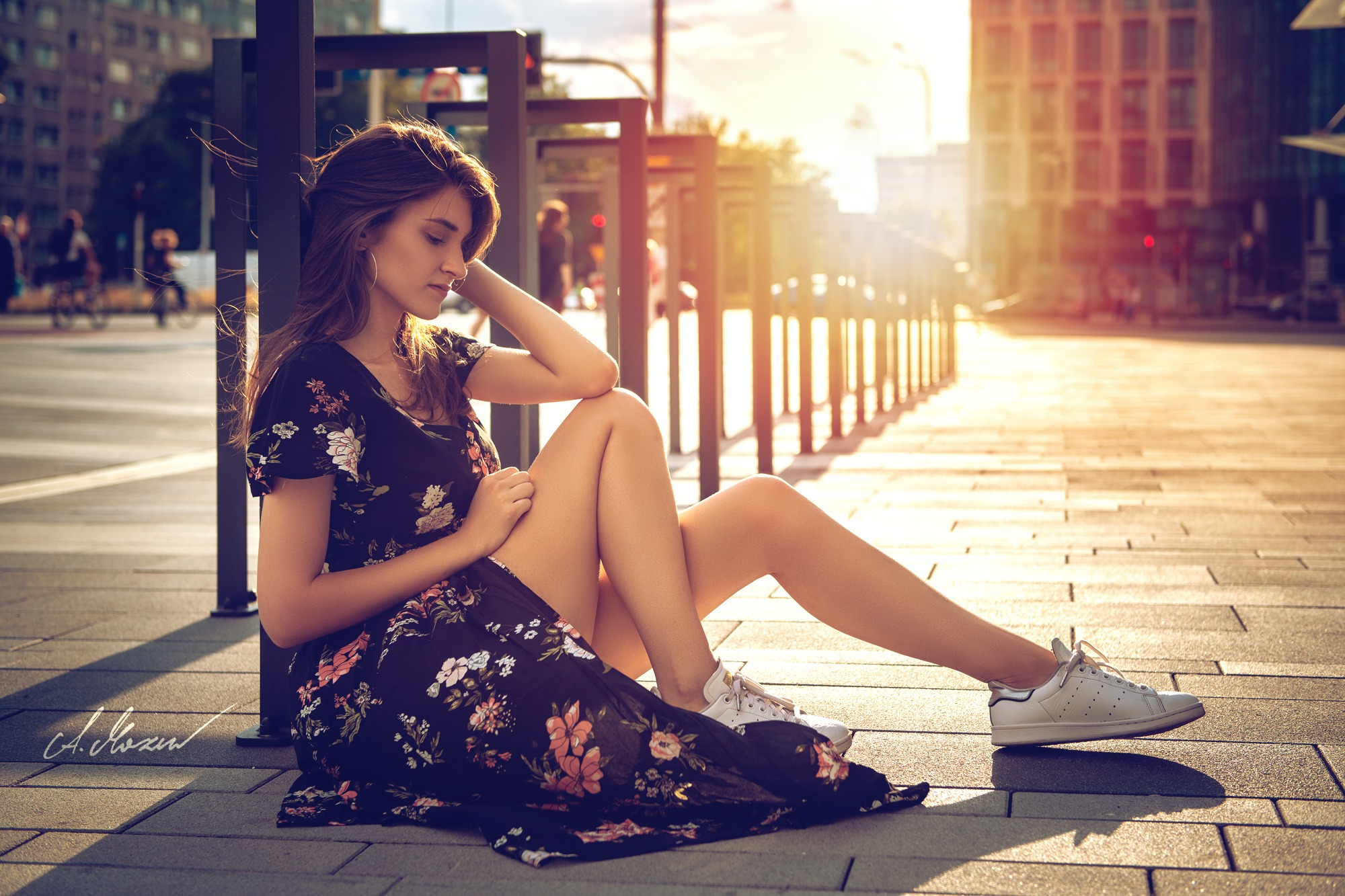 Młoda kobieta w kwiecistej sukience i białych sneakersach siedzi na chodniku w miejskim otoczeniu, opierając głowę o dłoń, z promieniami słońca w tle.