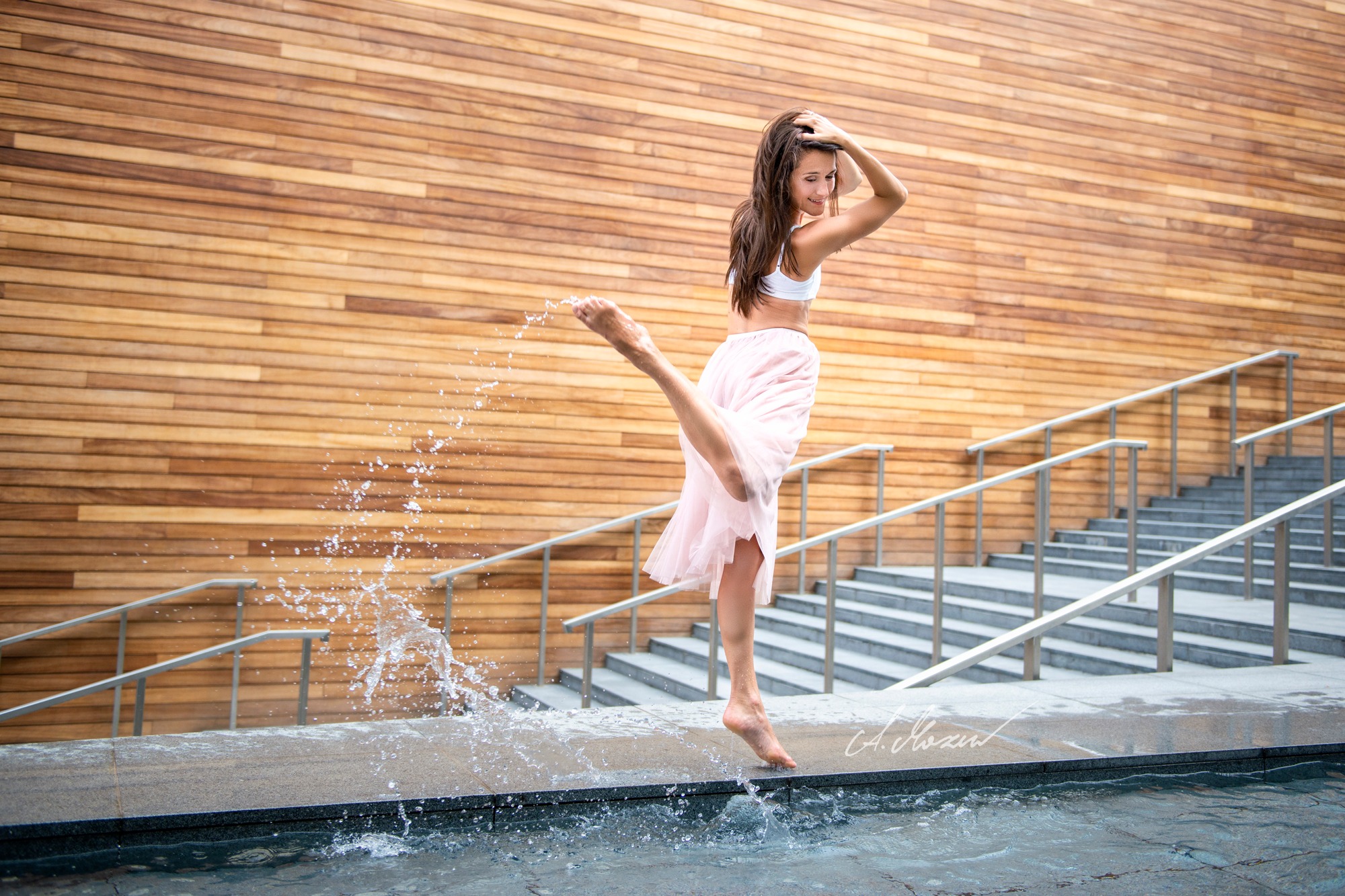 Kobieta w białym topie i różowej spódnicy tańczy na tle drewnianej ściany, unosząc nogę i rozpryskując wodę z fontanny.