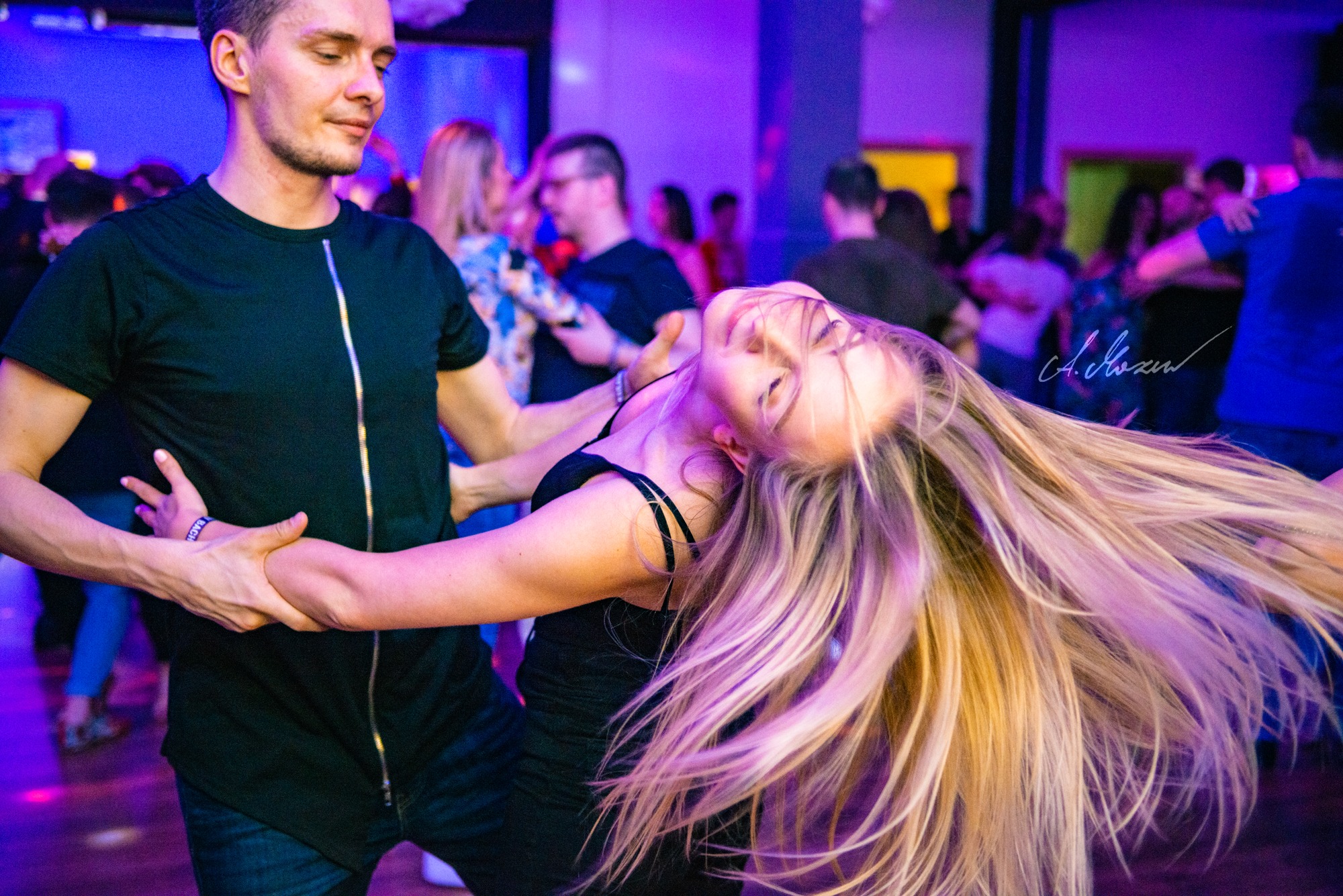 Para tańczy na parkiecie w fioletowym oświetleniu, kobieta z blond włosami odchylona do tyłu, mężczyzna w czarnej koszulce z suwakiem trzyma ją za ręce.